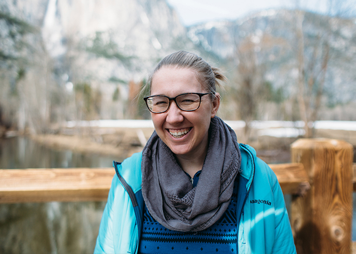 woman wearing glasses, a blue jacket, and a scarf laughing in front of mountain scenery